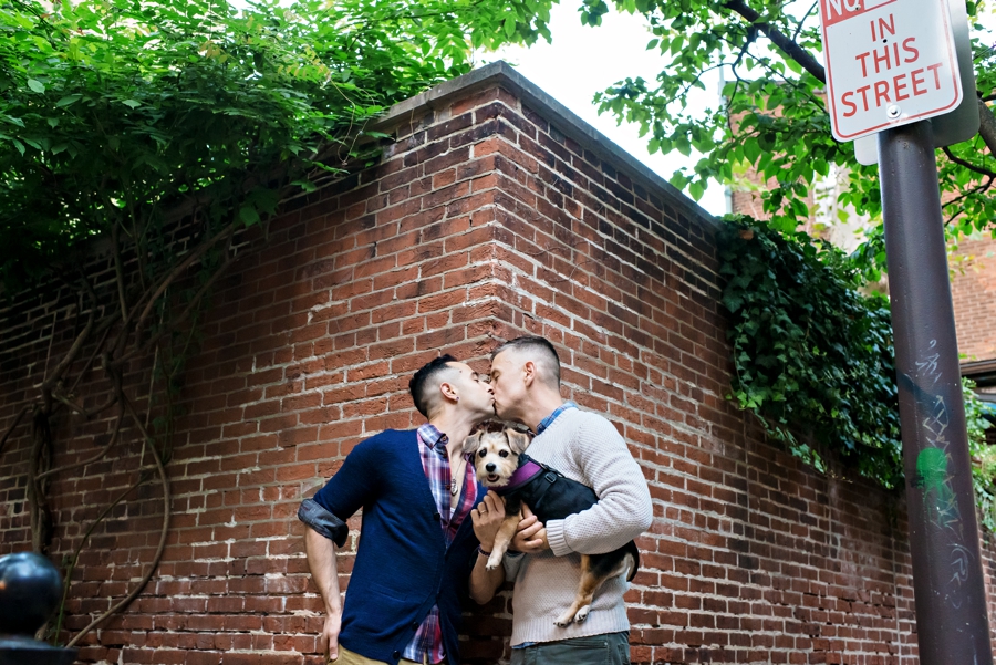 Two grooms kiss while holding their small dog along a brick wall in Philadelphia.